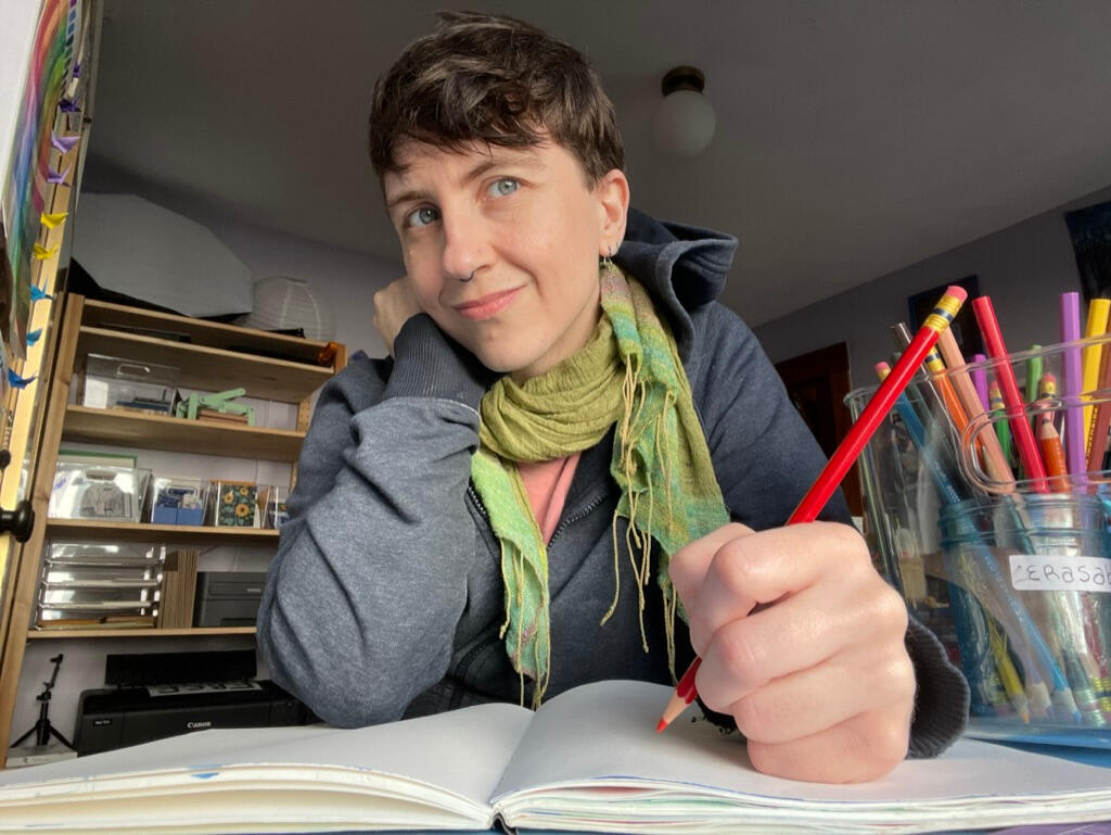 Portrait of Robin sitting at his desk holding a colored pencil to a blank sketchbook page. He is light skinned and androgynous, with short brown hair and blue eyes. He's wearing a hoodie and a green scarf.